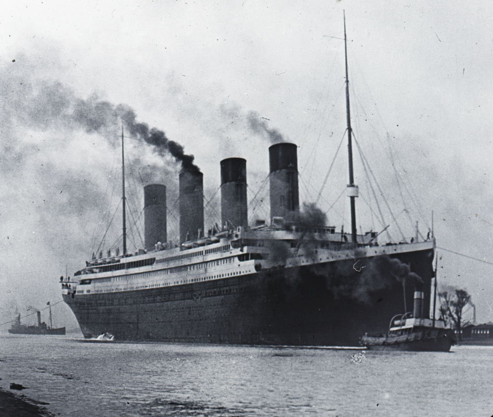 A very large ship with steam coming out of four columns, moving alongside two smaller boats.