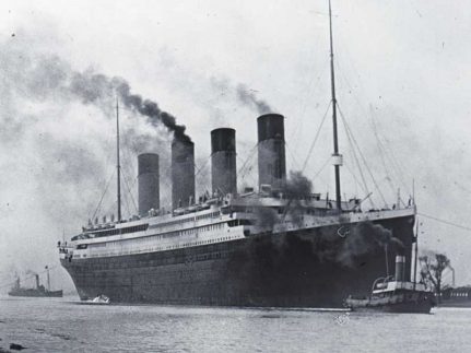 A very large ship with steam coming out of four columns, moving alongside two smaller boats.