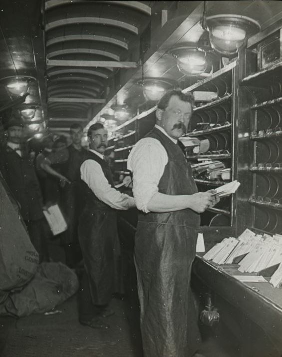 Men standing in front of sorting frames sorting mail, photographic lantern slide, early 20th Century