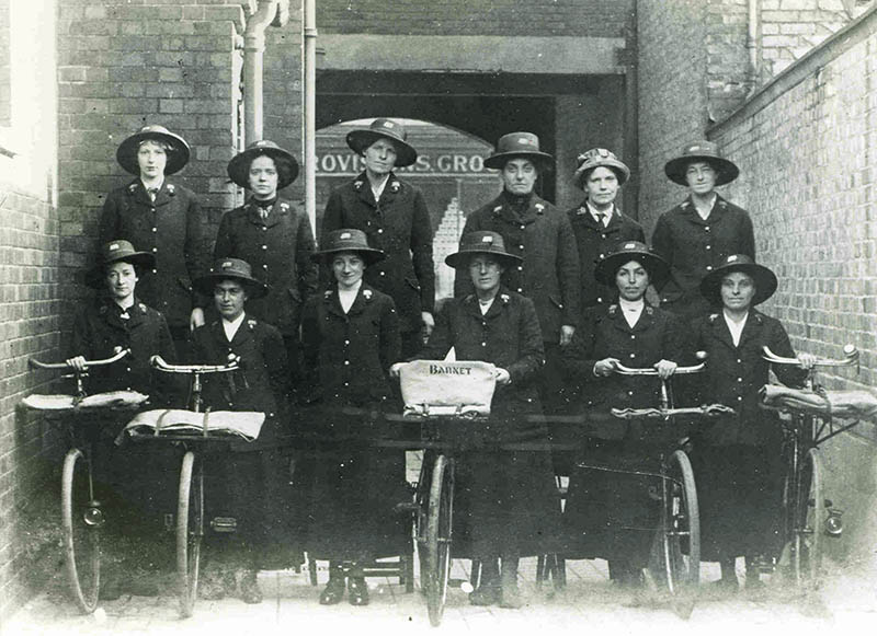 A staged photograph of postwomen wearing the Post Office's first uniform (a long coat and straw hat). Some of them hold bikes, one with a fabric basket labelled "Barnet".