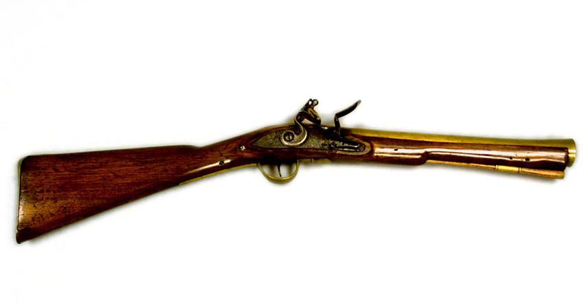 A Blunderbuss, 1788-1816. Believed to be of Post Office origin and used on Mail Coaches. Wooden, polished walnut stock and butt with decorative brass butt cap and mounts. Brass trigger guard protecting [iron] trigger. Brass 14 inch barrel with bore (or internal diameter) which widens towards the muzzle. Birmingham proof marks on barrel. Iron, non-decorated flintlock lock (firing mechanism) with flint present and mounted in a folded material [leather]; there is also a hinged bolt safety catch. No manufacturer's marking on lockplate. Non-original wooden ramrod with horn tip which slots into stock and ramrod pipes beneath the barrel.