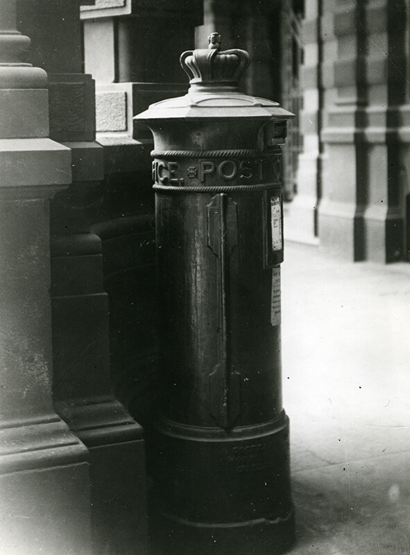 A pillar box for letters standing on the corner of a street, with a metal crown attached to the top of the box. The words 'Post Office' are on the pillar of the box.