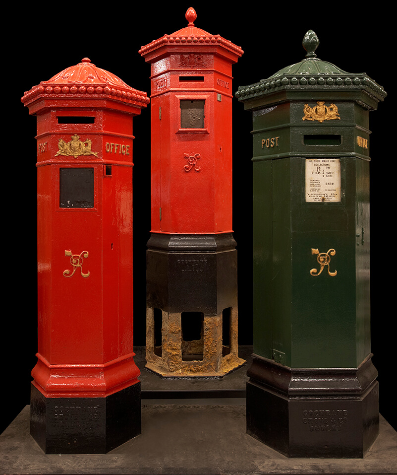 Three pillar boxes standing against a plain black background. Two boxes are red, and one is green.