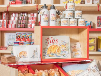 A colourful display of children's mugs, bottles, books, toy cats and stationery on a shop trolley.