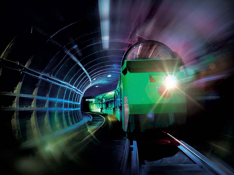 A photograph of a green miniature Mail Rail train travelling through an underground tunnel.