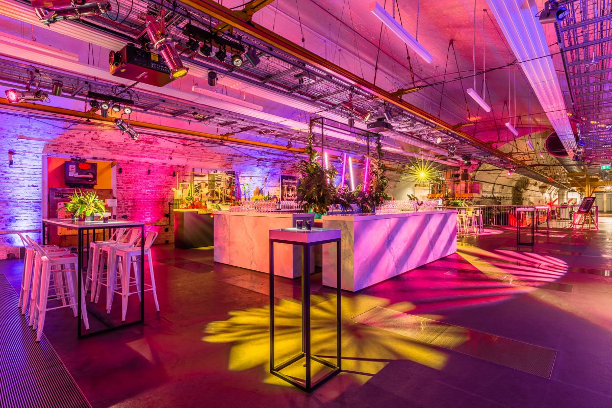 An underground venue room decorated with pink chairs, tables with flowers, and pink and yellow lighting. The Mail Rail tunnels are visible in the background.