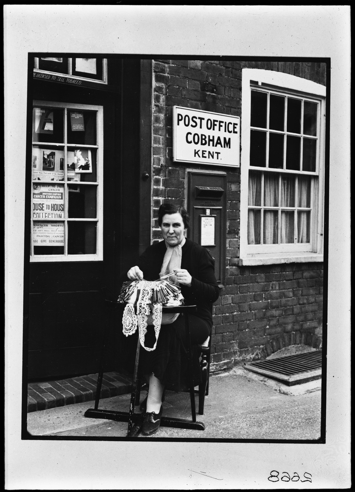 Black and white photograph of a women sat outside of Cobham Post Office making lace.