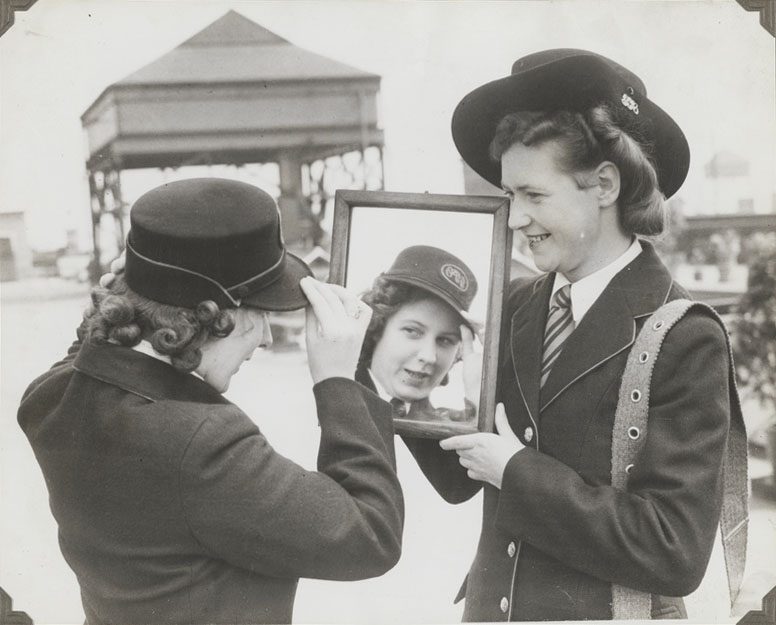 Black and white photograph of a women adjusting her postwomen hat in a mirror.