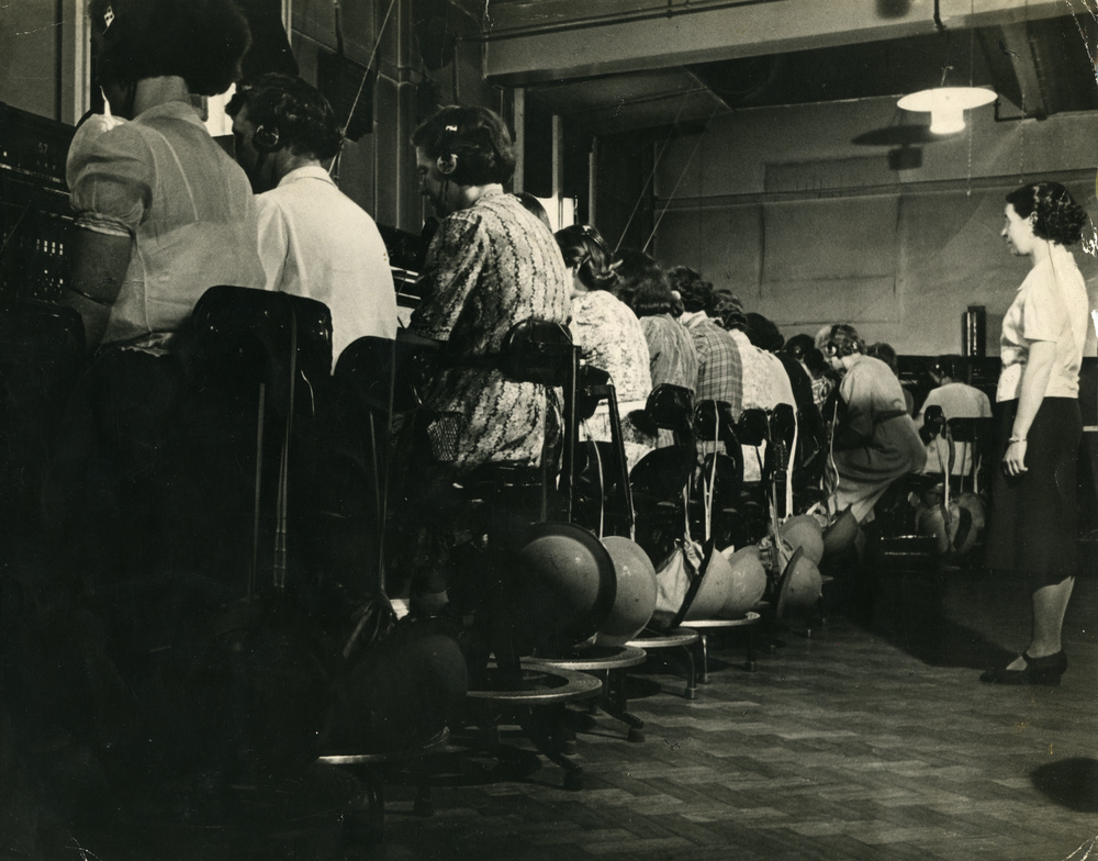 Black and white photograph of women sat in a line working the telephone exchange with a supervisor. 