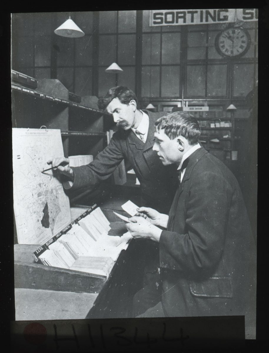Two postal workers at Mount Pleasant sorting office. One postal worker is sitting sorting letters, while the other stands beside him and points to a location on a map.