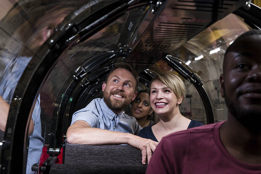 Adult visitors seated in pairs inside a miniature train carriage, smiling as they look out through the windows.