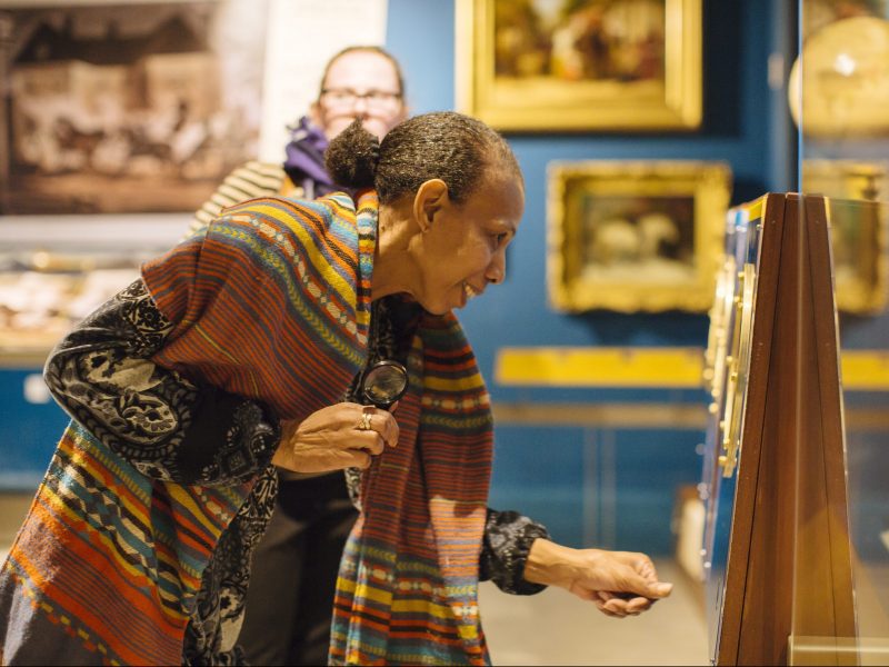 An older women wearing a striped multicoloured cardigan, looking at a wall display in the museum gallery.