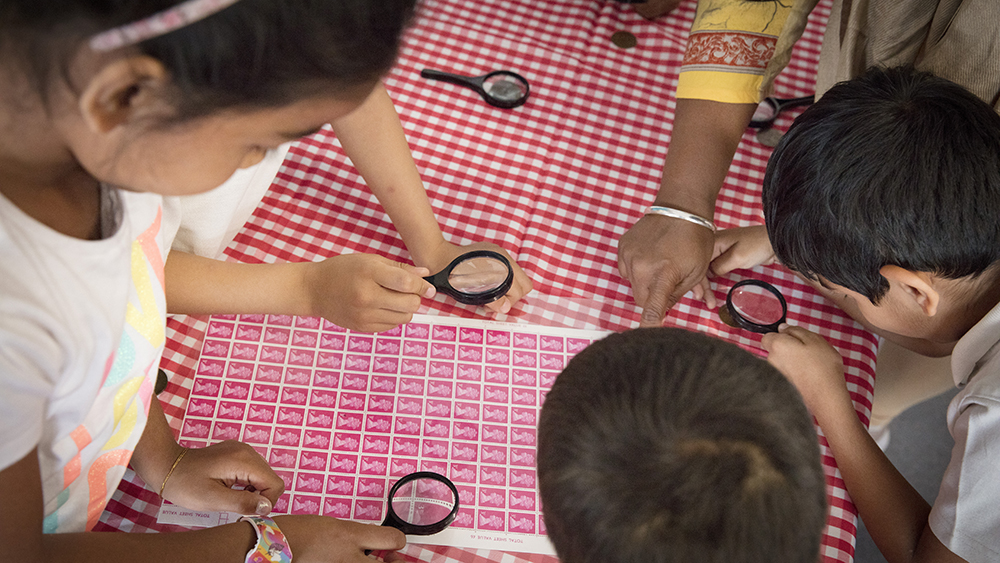 An image of children bent over a sheet of stamps using magnifying glasses to get a closer look. 