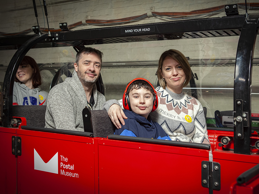 A child wearing ear defenders looks at the camara and smiles. He and his family sit on the Mail Rail ready for departure.