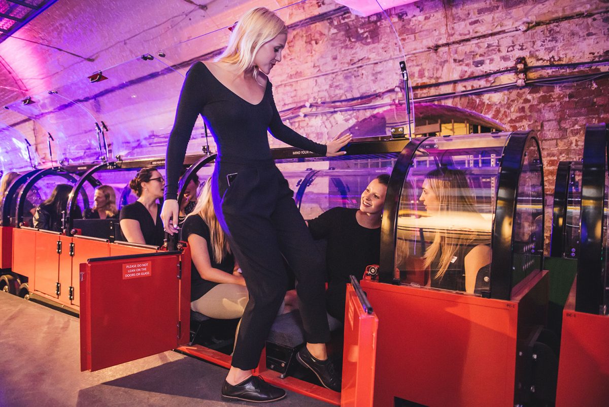 A smiling woman is stepping into a red miniature train. She has blonde hair and is wearing a black top and trousers. Other passengers on the train are smiling.