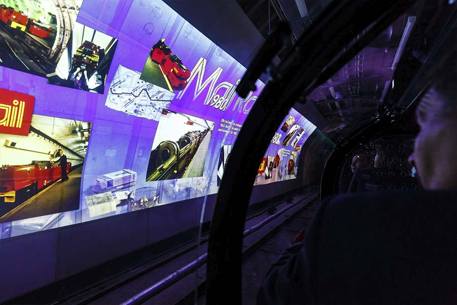 A slideshow plays on the walls of a train platform. The slideshow is projecting photographs of old trains, with the words 'Mail Rail' on display. Dark tunnels are visible at the end of the platform.