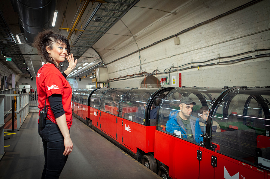 A woman with brown hair and a red museum uniform waves at a departing miniature red train. A father and son are sitting in one of the carriages, waiting for the ride to depart.