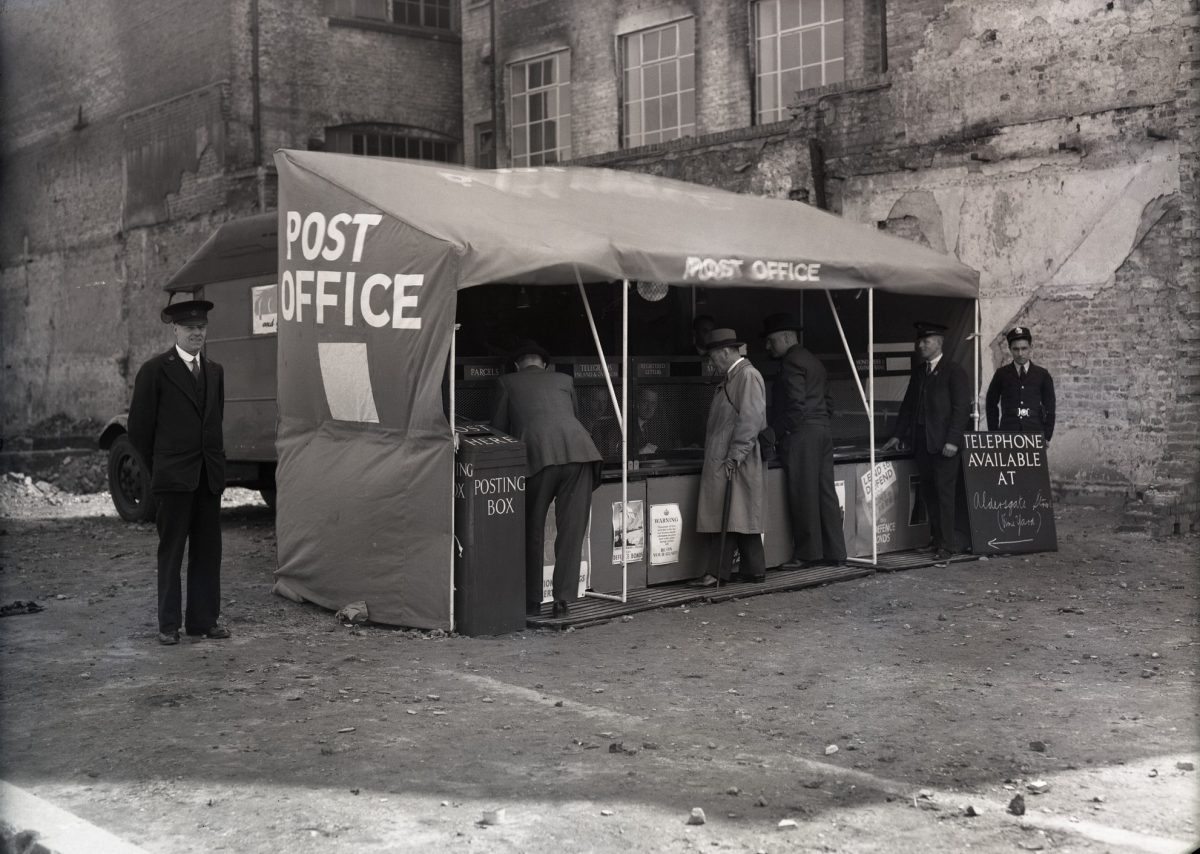 black and white photograph of a tent with a sign ‘Post Office’ and temporary post office counters inside. There is a temporary ‘posting box’ and blackboard advertising the availability of telephone services. There are men queuing to use the Post Office and postal staff in uniform nearby.