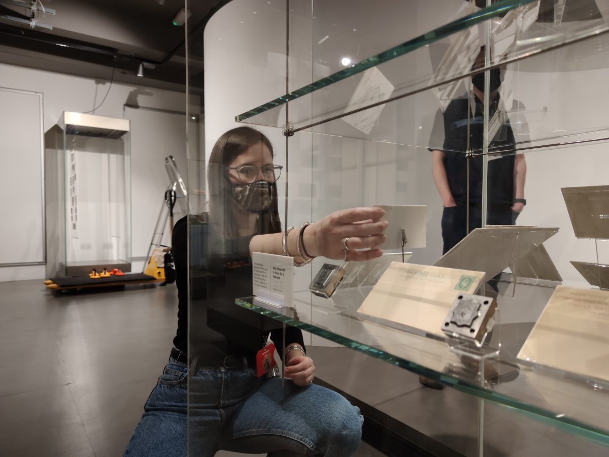 Image of a woman wearing a mask placing a item caption onto a shelf in a display case. 