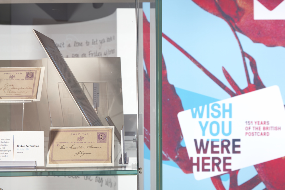 Close up image of postcards in a case with the 'Wish You Were Here' poster in the background. 