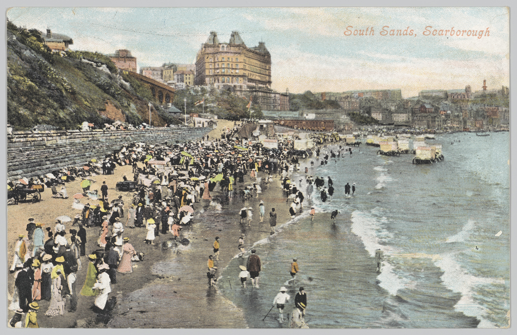 Postcard depicting many people on a beach with a large building in the background. 