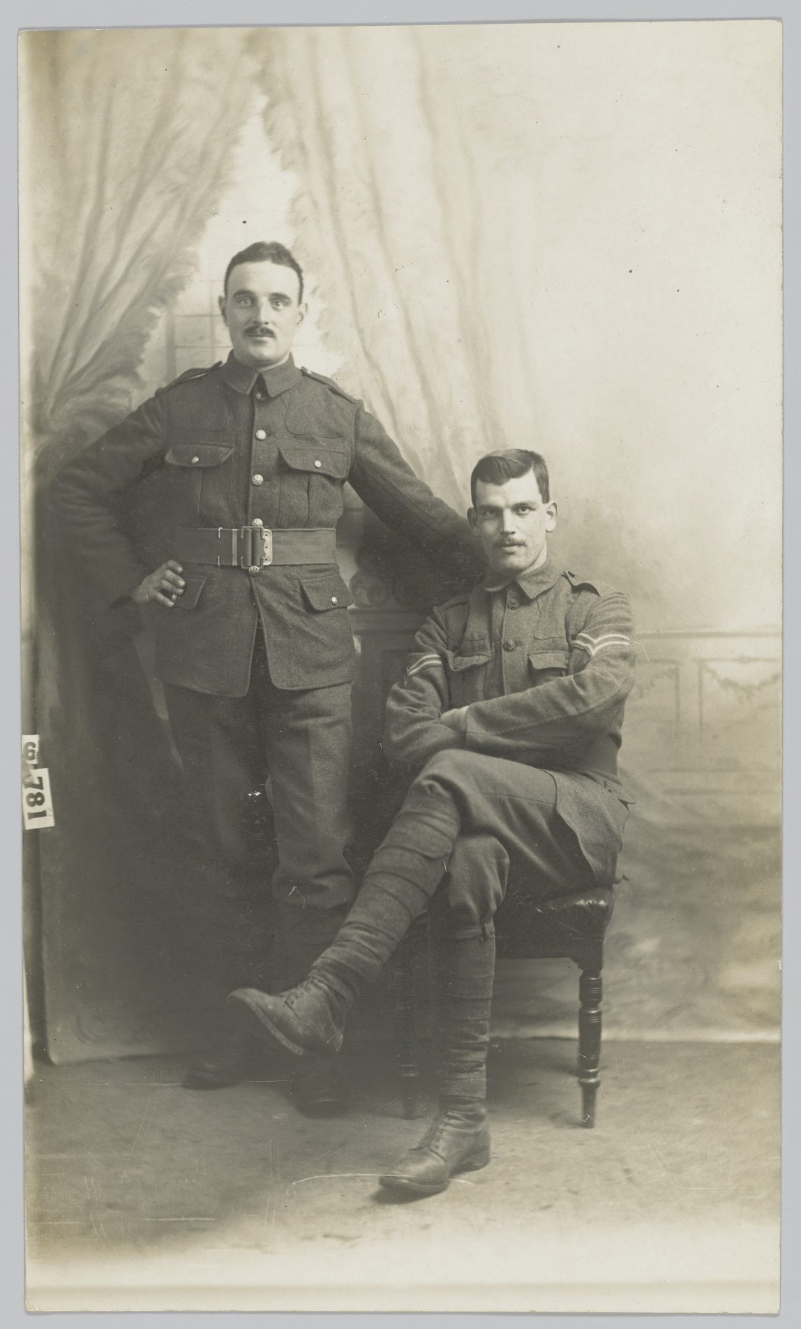 Black and white photo of a soldier sitting and another standing next to him with his arm on the back of the chair.