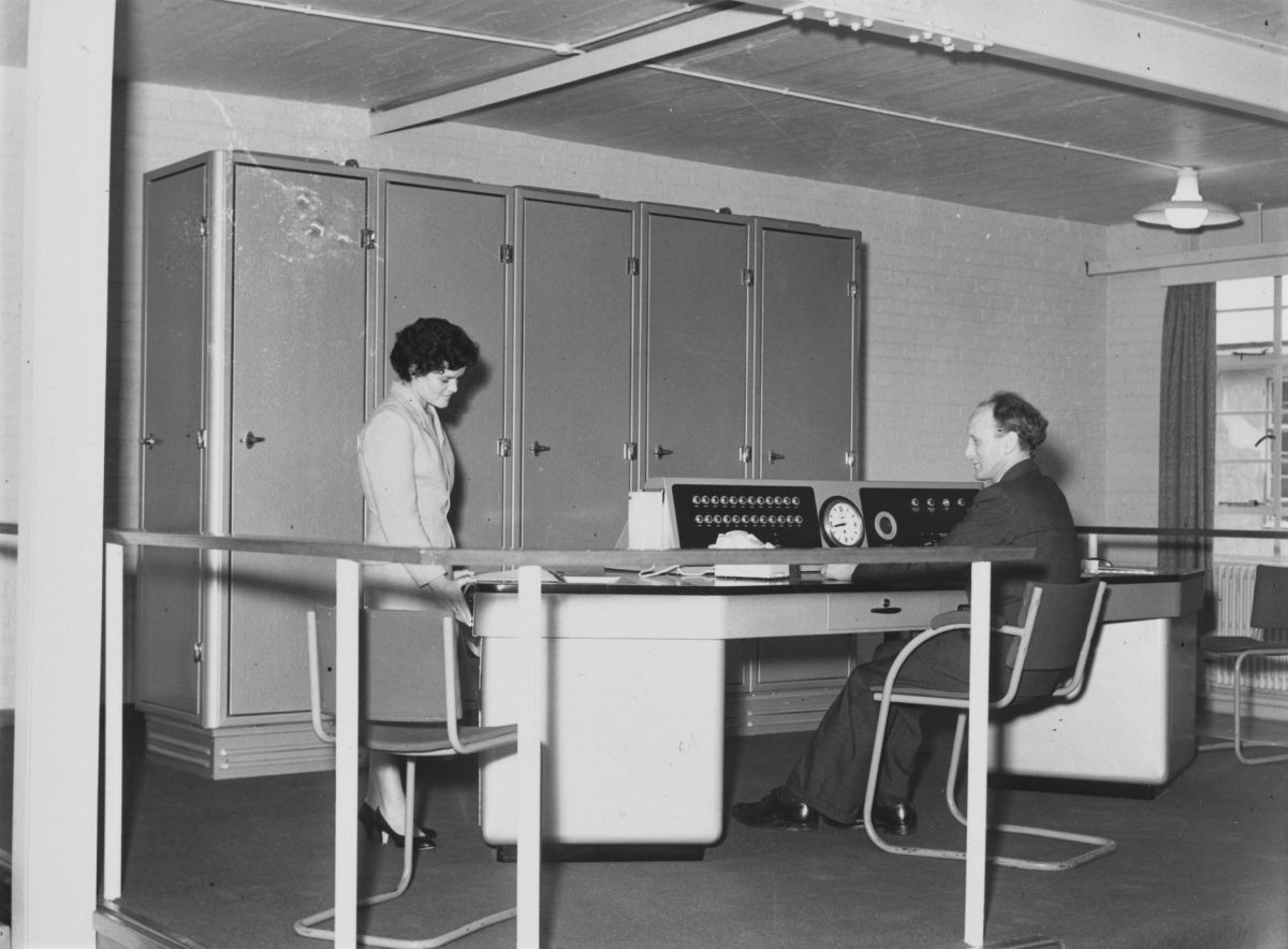 Black and white photograph of a white man sat at a table with a machine on the table. There is a white woman standing on the other side of the table. Along the wall are tall filing cupboards.