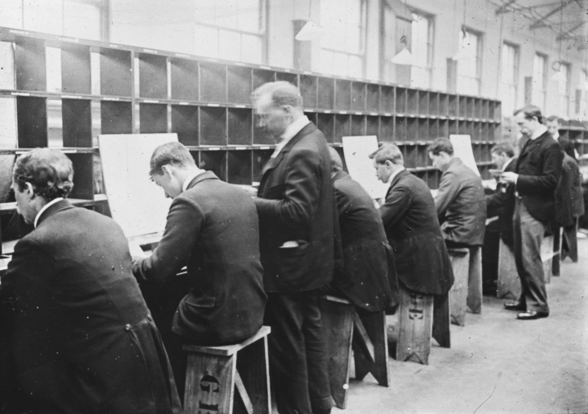 A line of men in black uniform jackets, seated behind a long row of shelves.