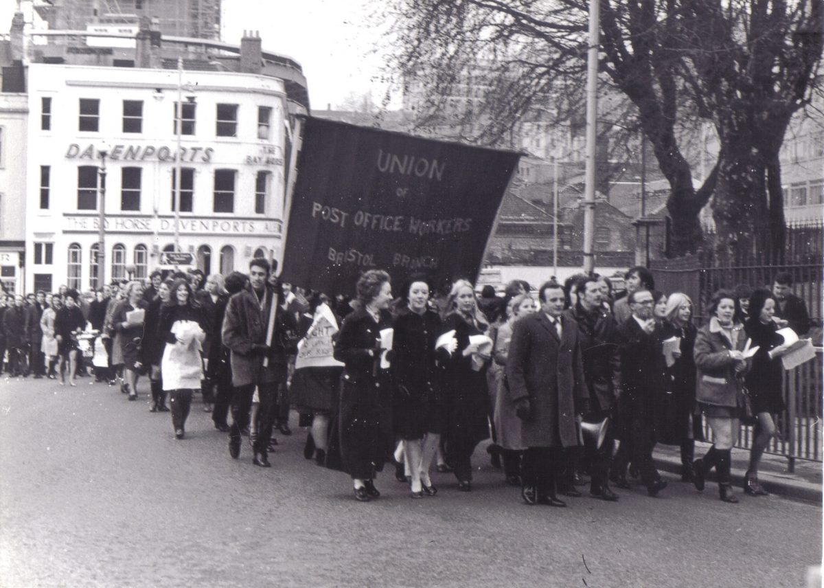 A black and white photograph of a group of people close together marching along a street. They are holding a banner with text ‘Union of Post Office Workers Bristol Branch’. To the right of the image are iron railings and in the background there is the sign for a shop called ‘Davenports’.