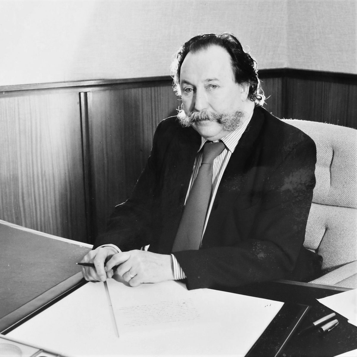 Black and white photograph of an older white male sat at a desk. The desk and person are at an angle to the camera. The man’s dark hair is receding, and he has a handlebar moustache. He is wearing a dark suit, white striped shirt, and a pale tie.