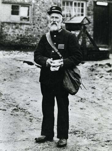 A black and white photograph of a man in an old postman's uniform, with th four white horizontal stripes on the left side of the jacket.