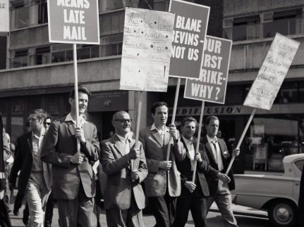 A black and white photo of six men carrying plaques with 'Blame Nevins not us' on them.