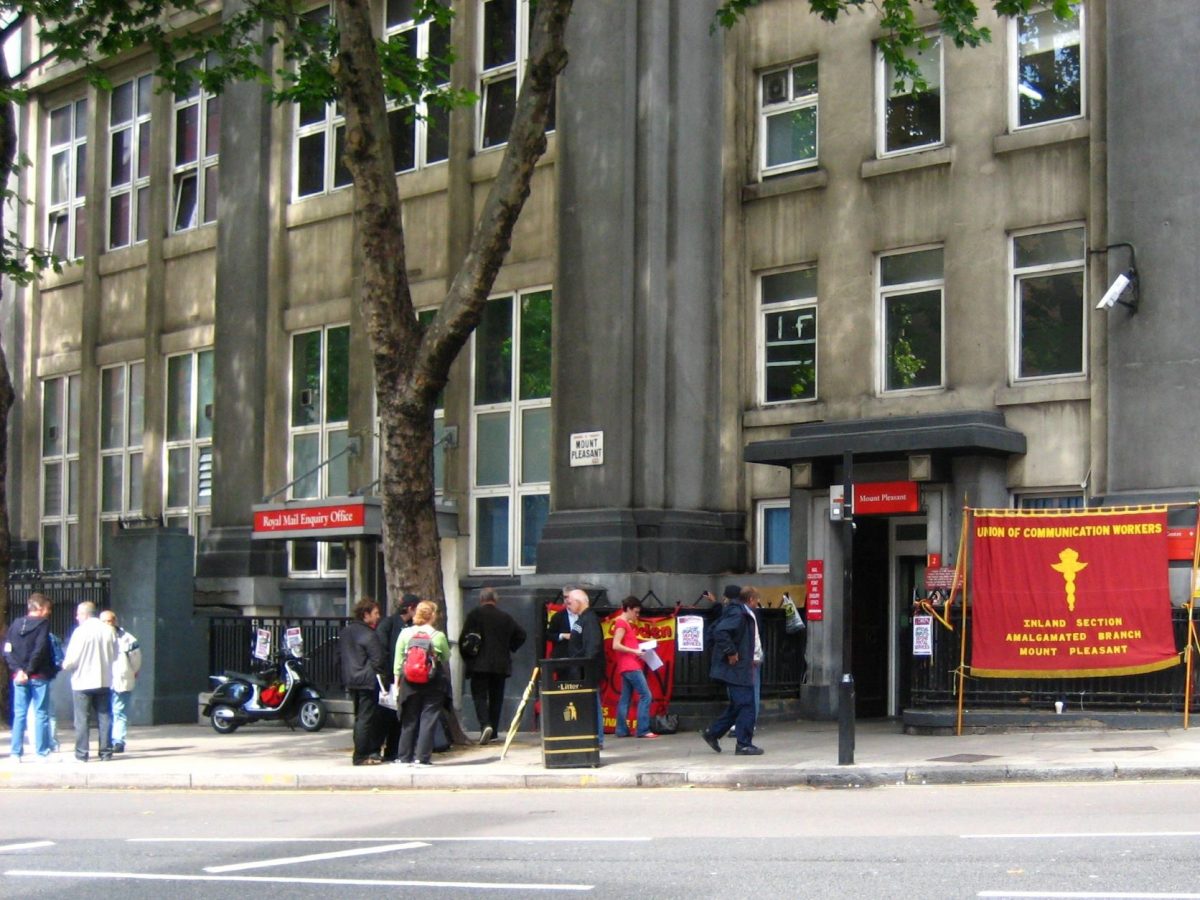 A coloured photo showing workers at a picket line outside a Royal Mail building. On display there is a large red banner saying the words: 'Union of Communication Workers.'