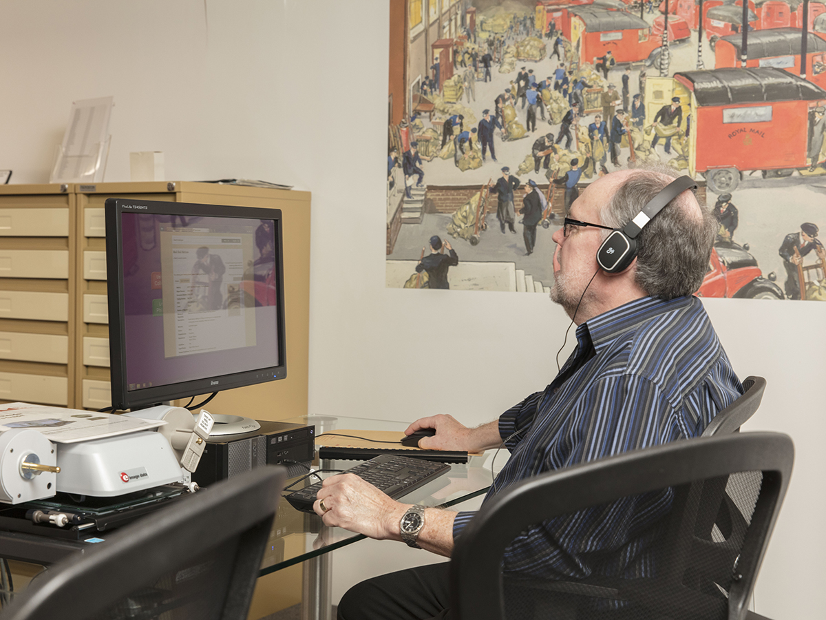 A middle aged man wearing glasses sits at a glass desk, using a computer. One hand is on the mouse and the other sits next to the keyboard. He sits comfortably in a desk chair.