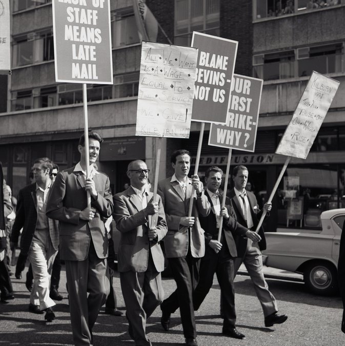 A black and white photo of six men carrying plaques with 'Blame Nevins not us' on them.