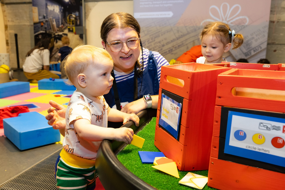 A Post and Play facilitator helps a very young boy put mini post in bright red postboxes. The child wears a top with tigers on it.