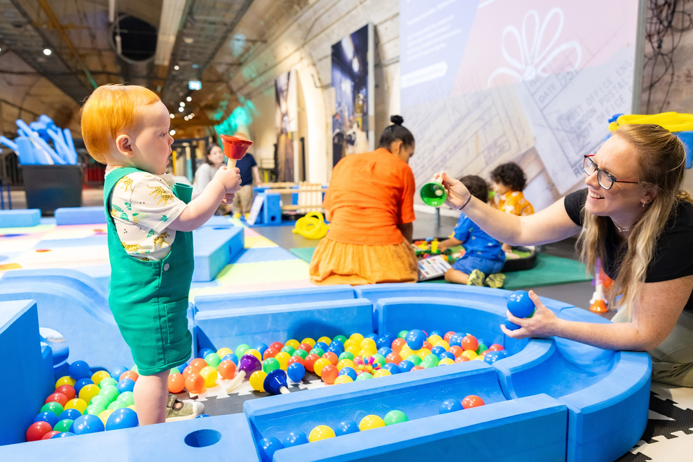 A toddler wearing bright green dungarees in a blue ball pit.