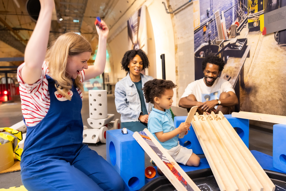 Rosie celebrates with a toddler as he successfully pushes a miniature car down a ramp. His parents look very proud of him.