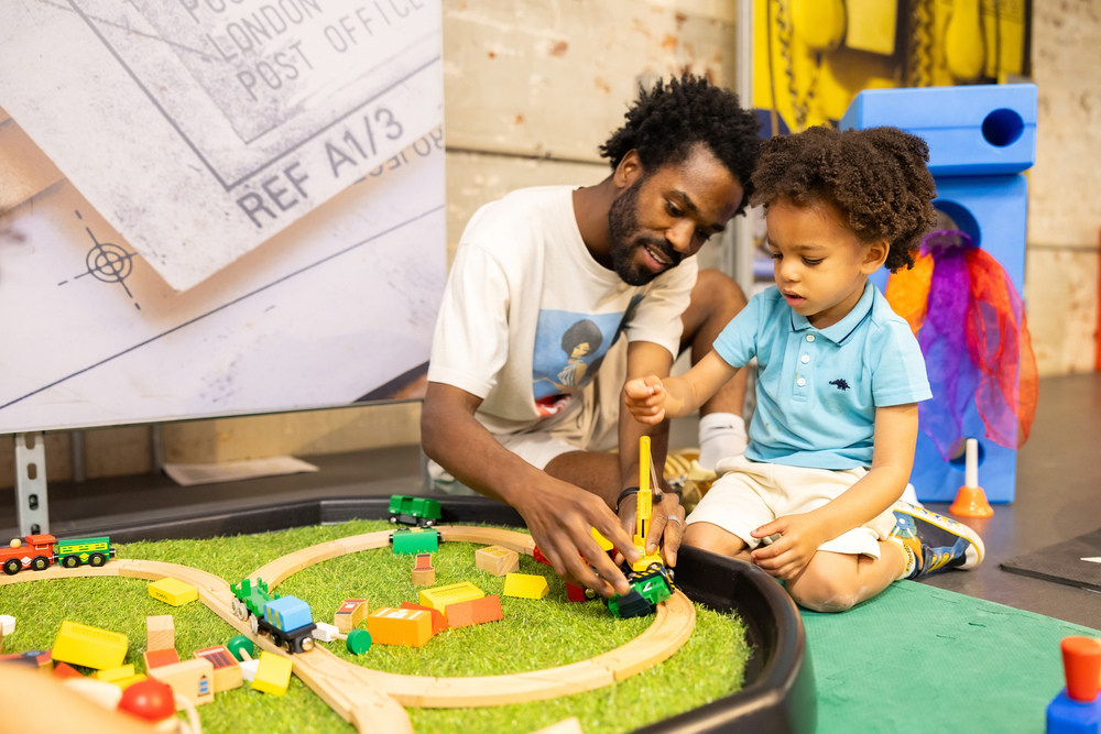 A man plays with his young son on a mini railway track. They are pushing miniature trains around the wooden track.