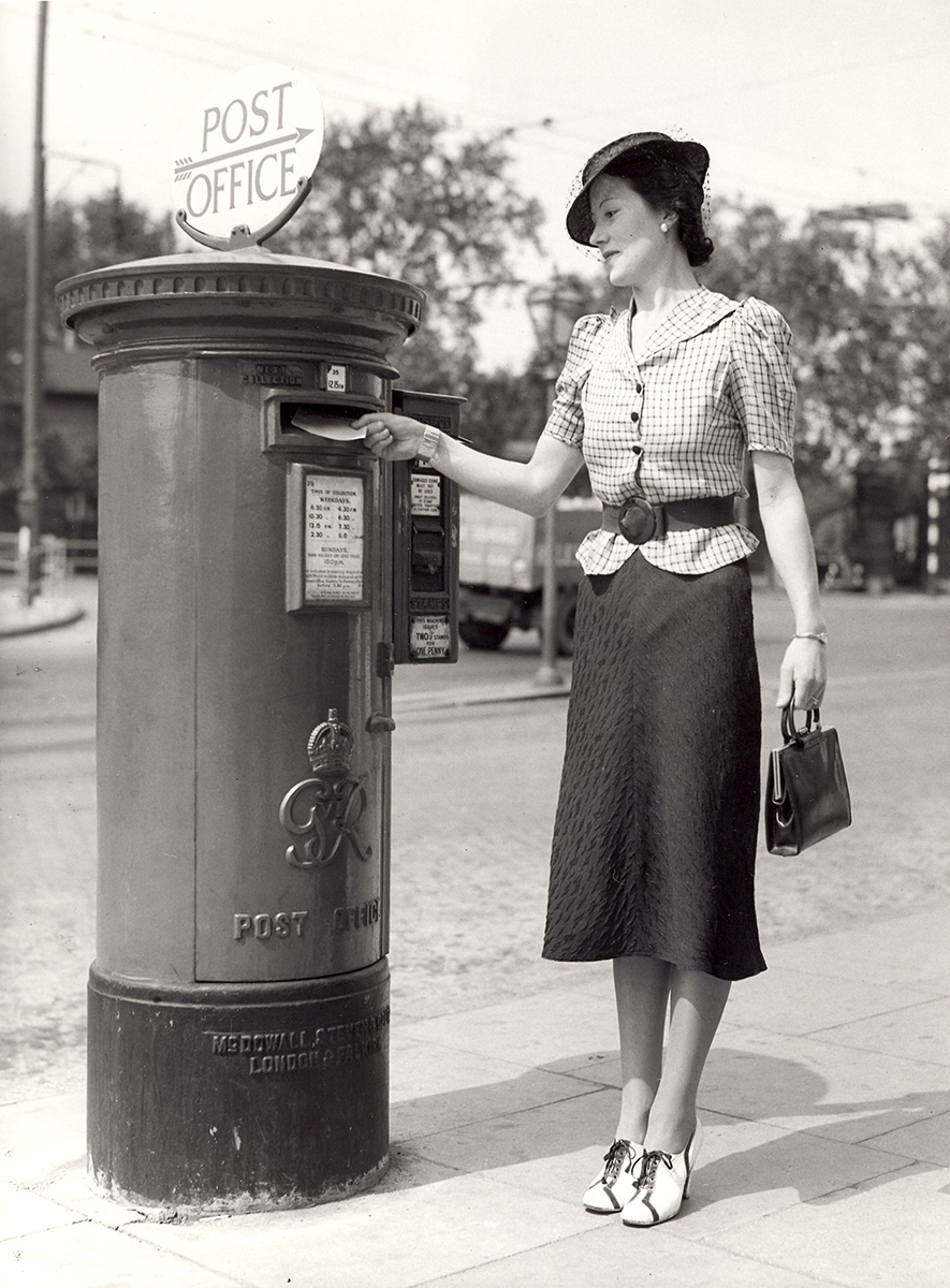 Black and white photo showing a King George VI pillar box. A lady posts a letter into a King George VI Pillar Box. The pillar box has a stamp vending machine attached to its side, and a sign on its top which points towards the nearest Post Office.