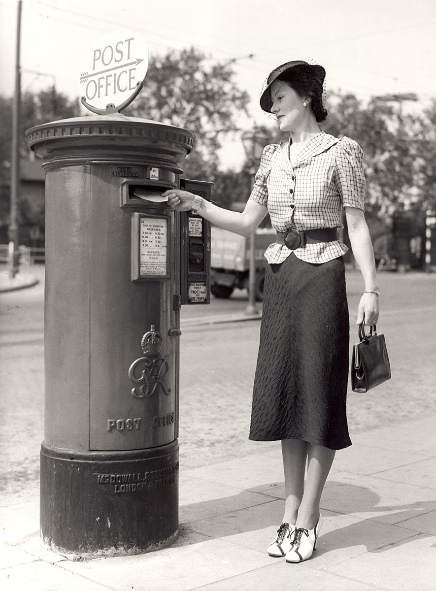 A lady posts a letter into a King George VI Pillar Box. The pillar box has a stamp vending machine attached to its side, and a sign on its top with a pointing arrow and the words 'Post Office'.