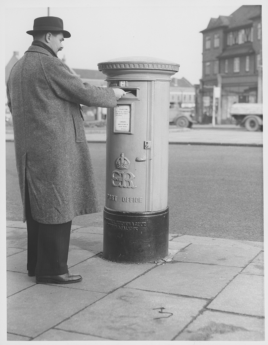 A man in a long coat and black trousers posting a letter into a pillar box with 'EVIIIR' cypher on the front.