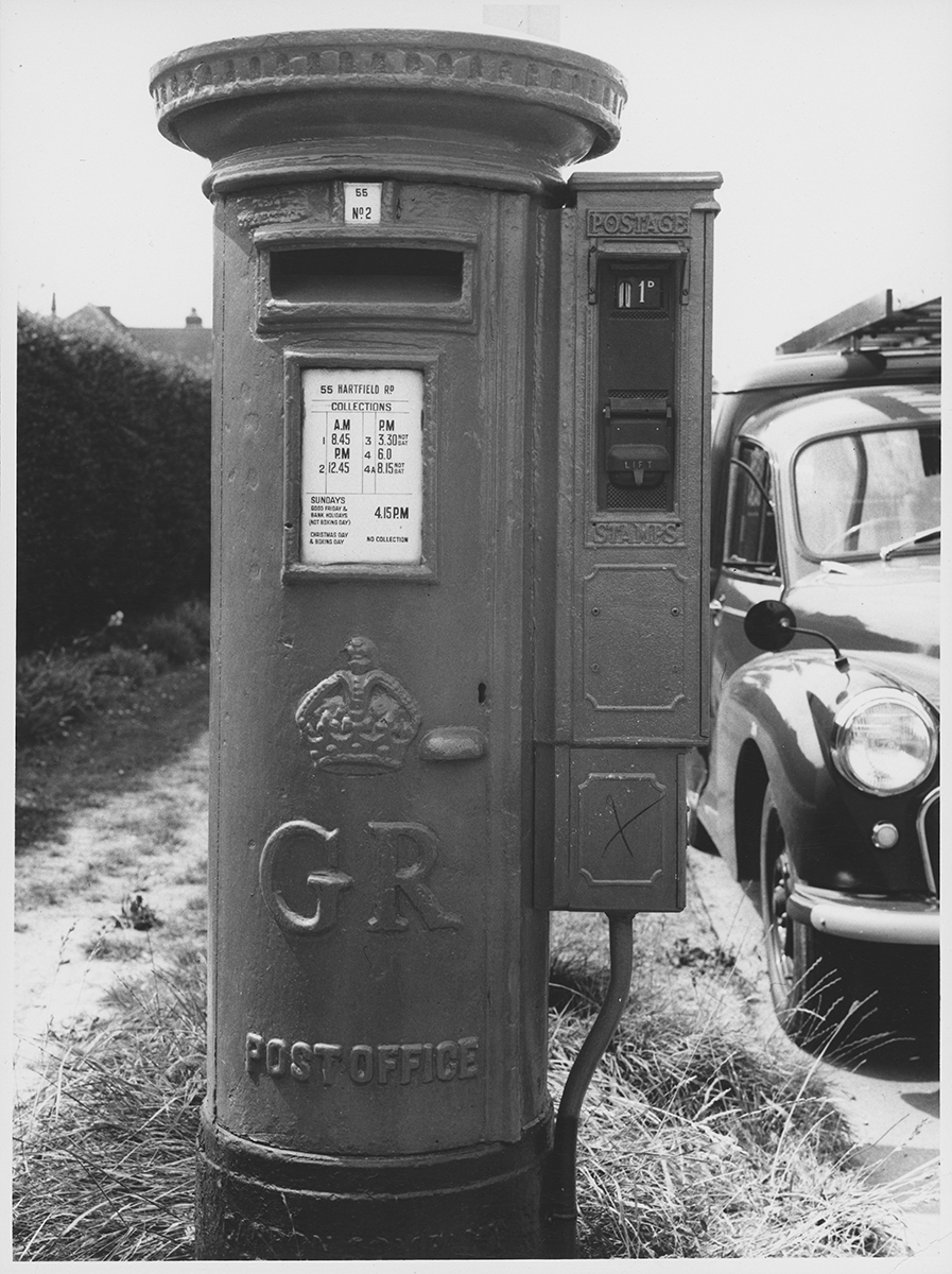 Photograph. Image orientation: portrait. Black and white. Exterior of a GR pillar box with stamp vending machine attached.