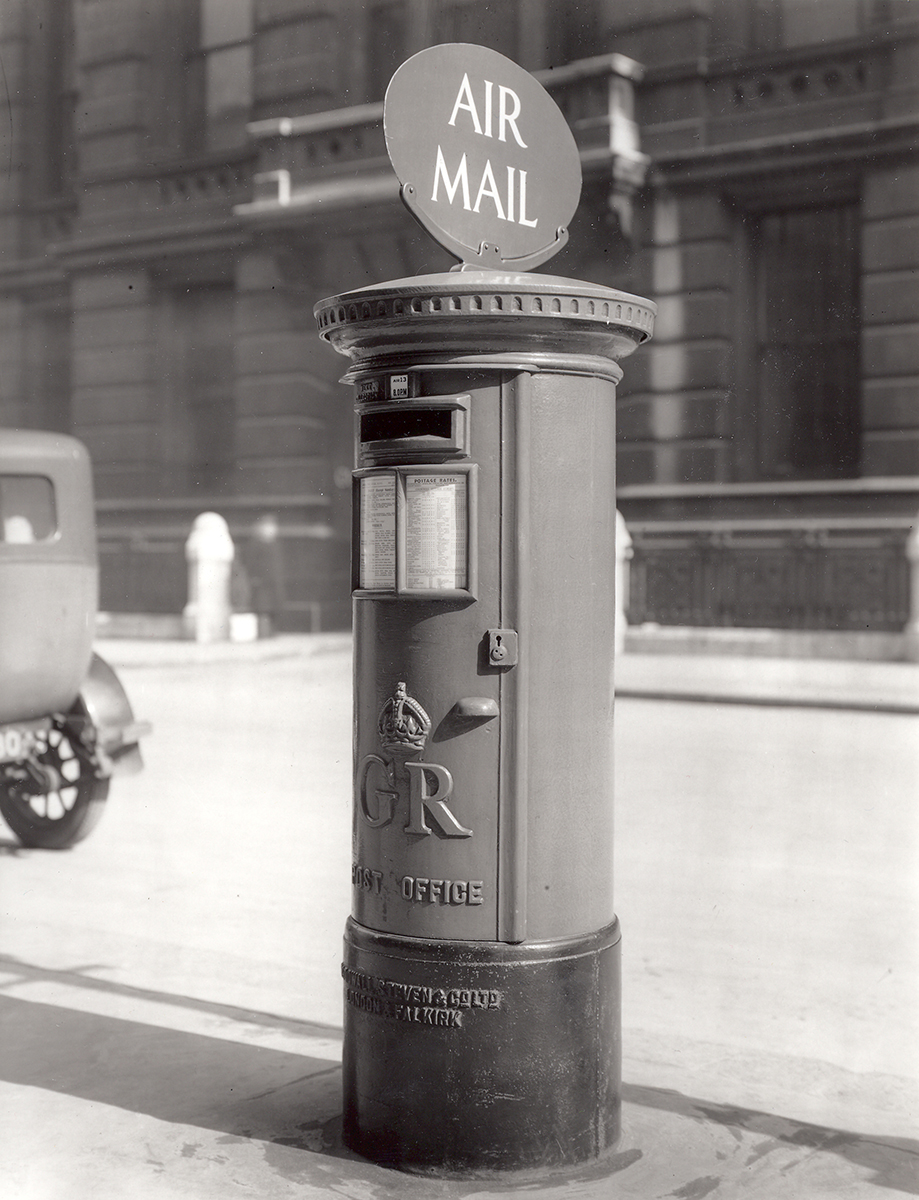 An Air Mail pillar box in the street, with a sign on top saying 'Air Mail'.