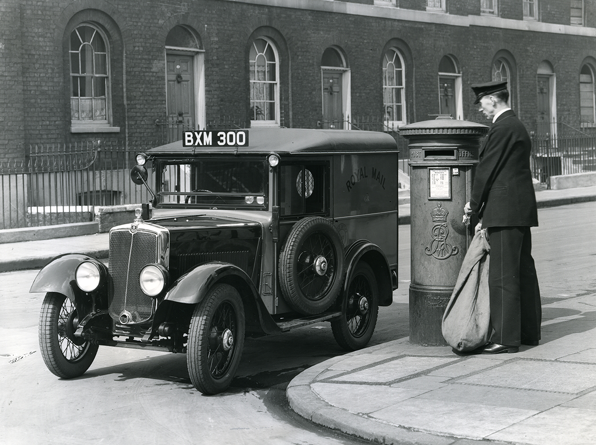 A postman collecting a sack of mail from a pillar box, standing in front of a Royal Mail van, registration number 'BXM 300'.