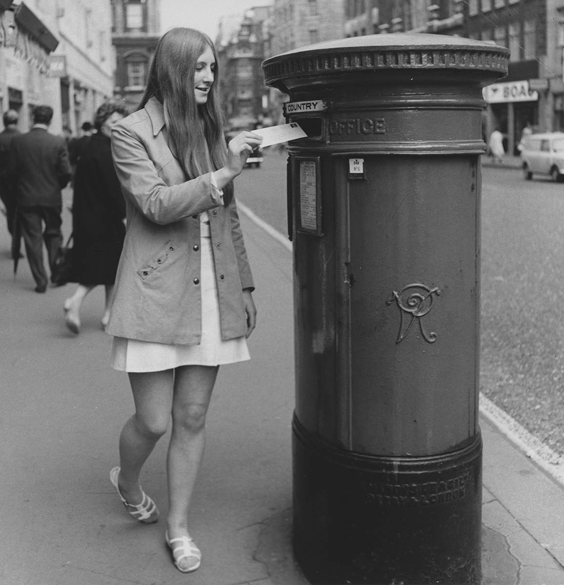 Photograph. Image orientation: square. Black and white. A woman posting a letter into a pillar box, in an unidentified London street.