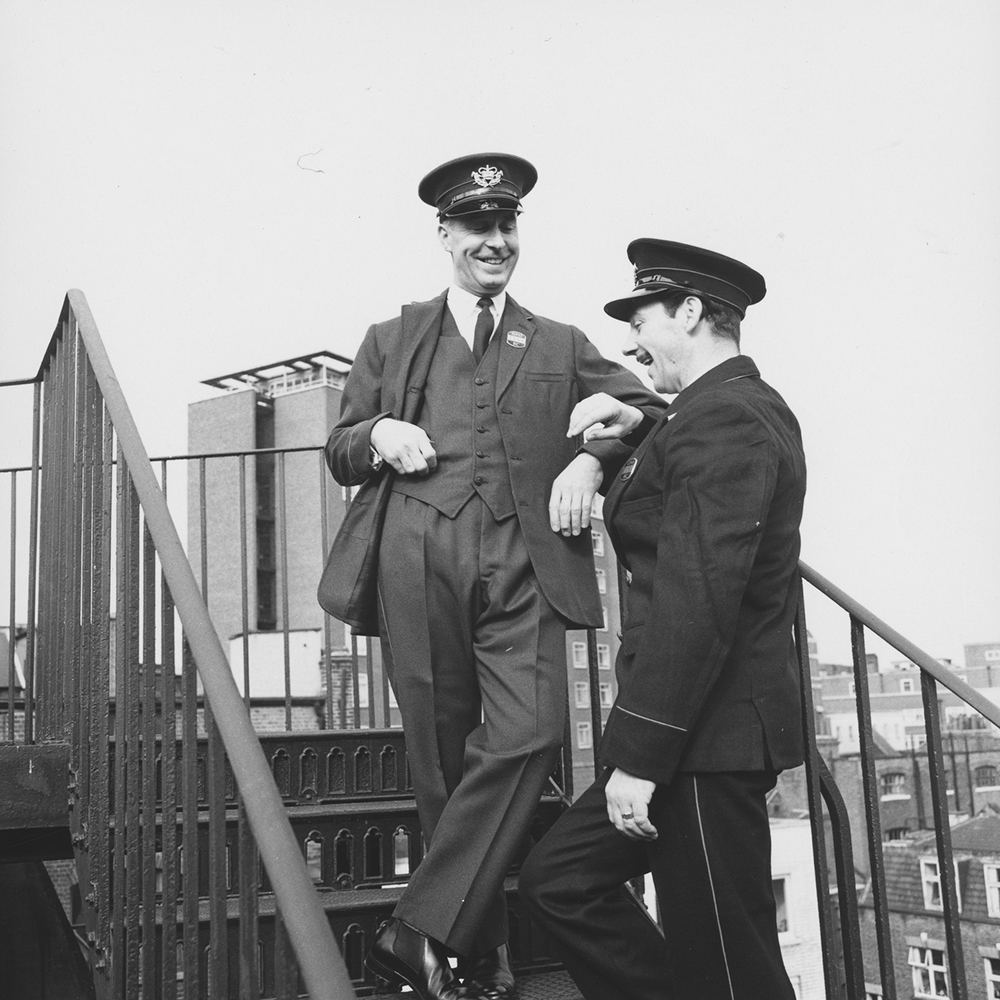 Photograph. Image orientation: square. Black and white. Two postmen standing on an outdoor stairwell. Photograph probably taken in London.