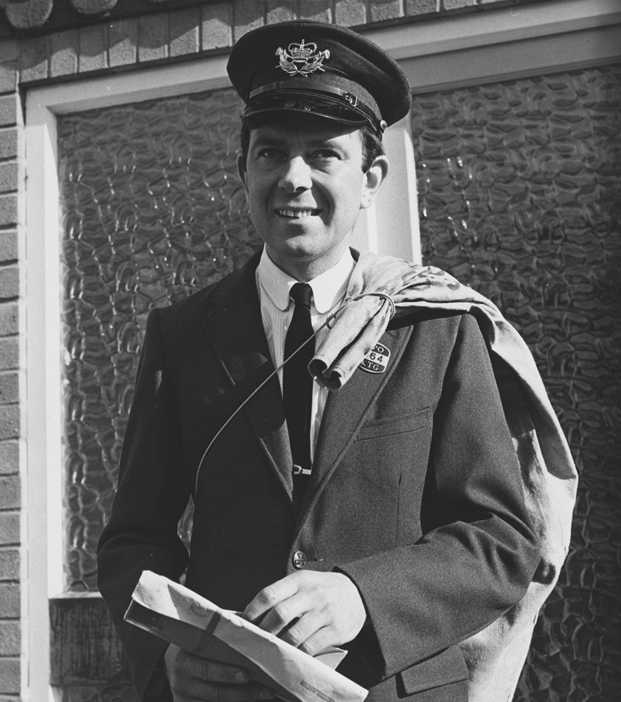 Photograph. Image orientation: square. Black and white. Postman on delivery in a residential area, holding letters.