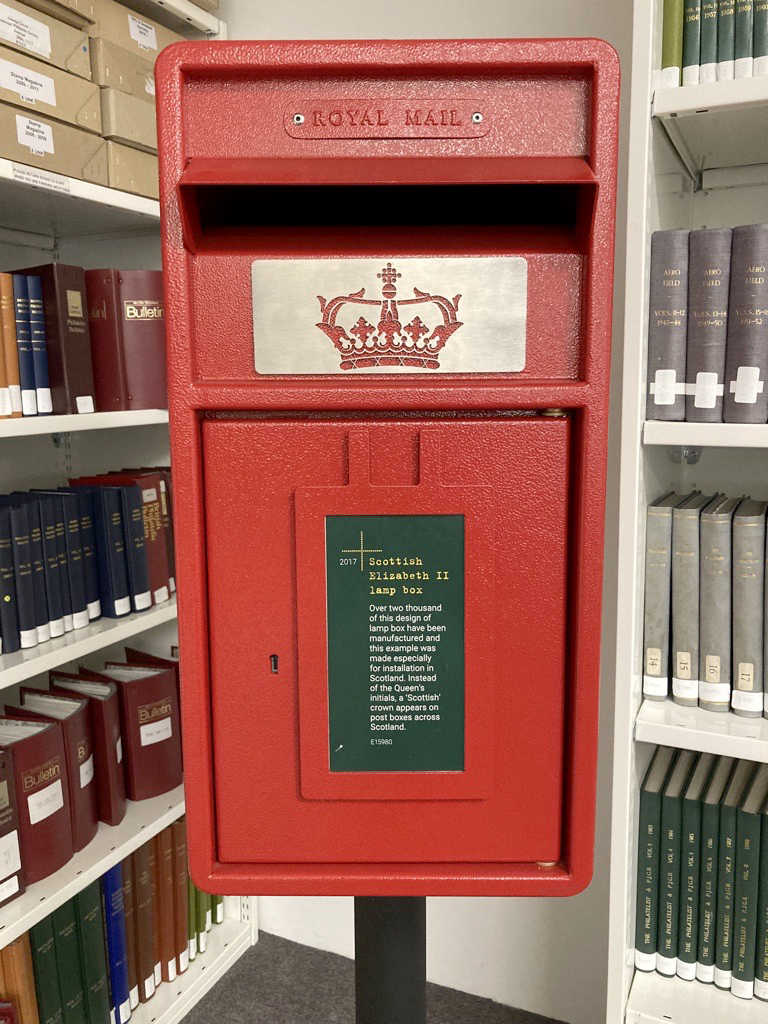 A coloured photo of a small red post box on a black pillar, showing the Scottish crown cypher below the letter aperture.