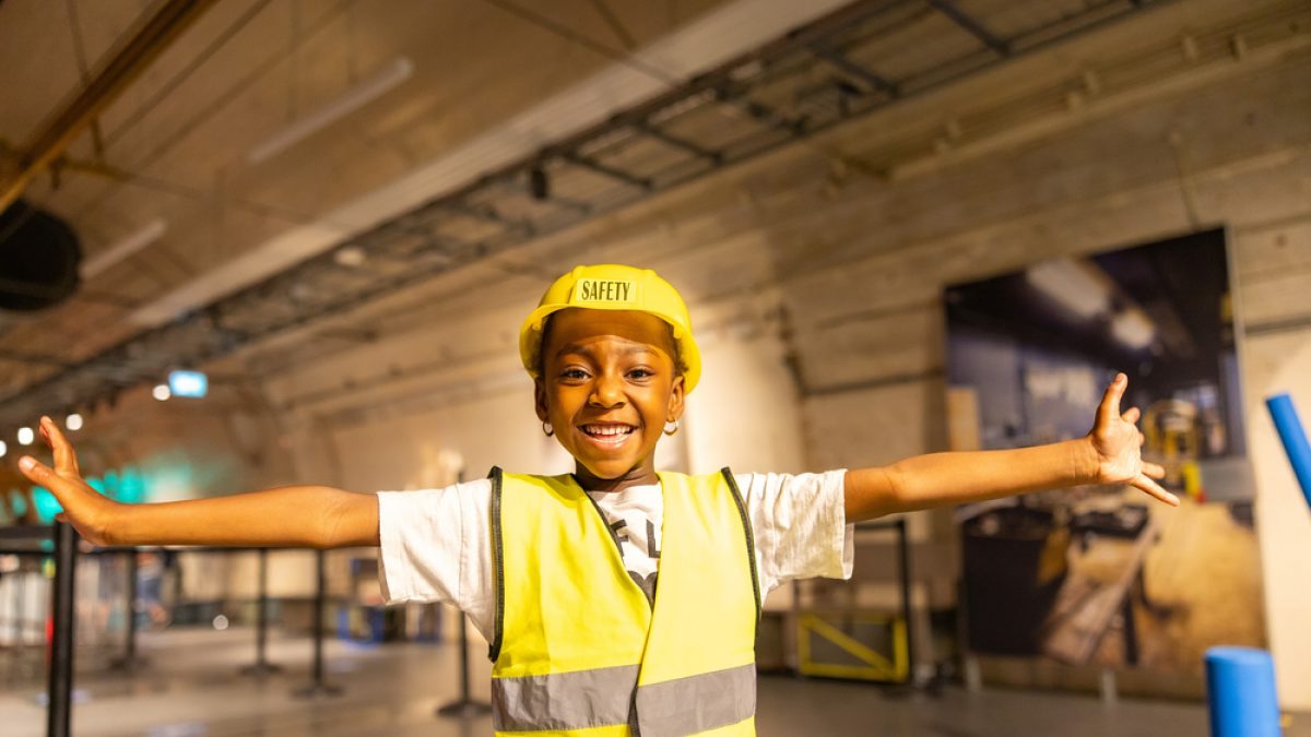 A girl wearing a high-vis vest and hard hat opens her arms and looks at the camara with big smile